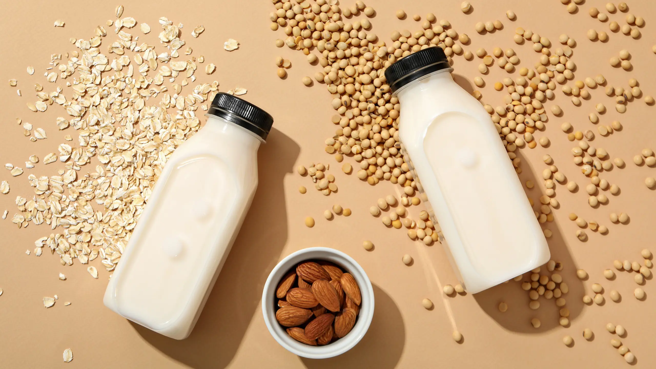 Bottles of milk, nuts in bowl, oatmeal and soy beans on beige background, top view