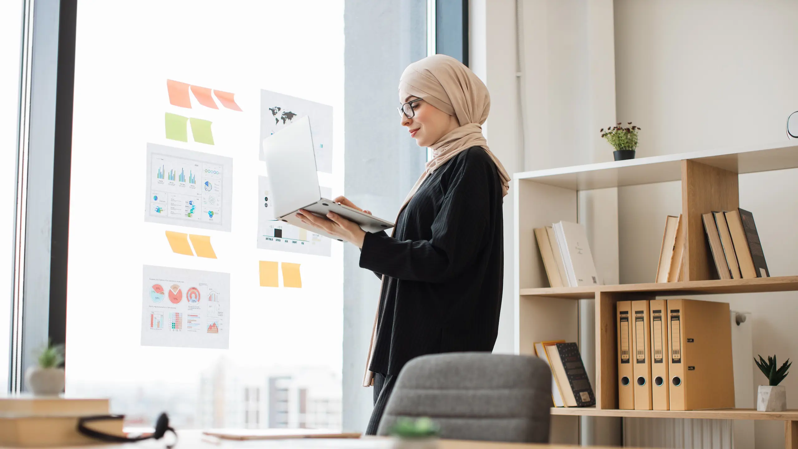 Thoughtful female in hijab typing on portable computer in hand while standing near glass window with graphic materials. Proficient arabian manager gaining better insight about new project at work.